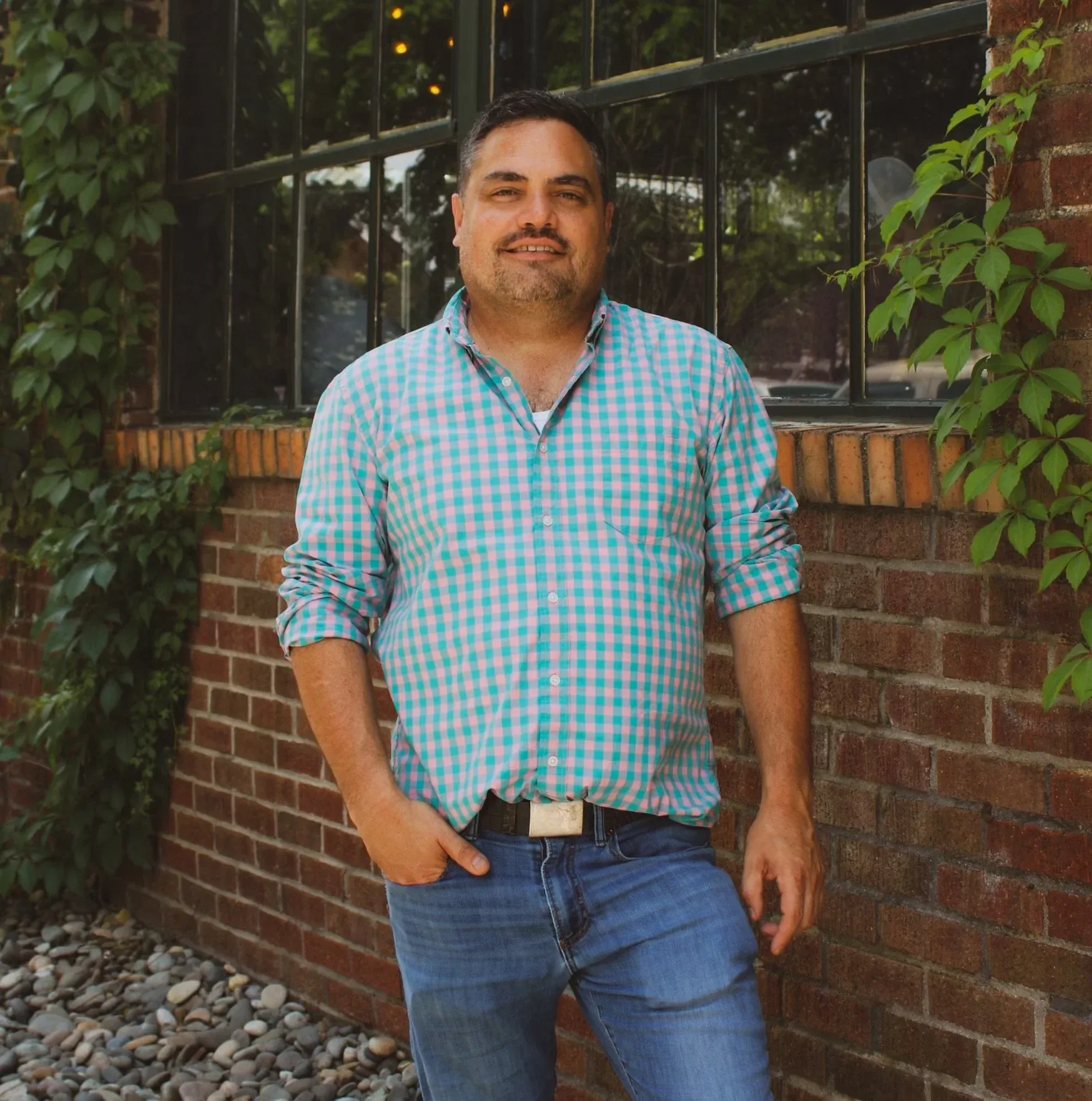 Chris Acosta outdoors against a brick wall with ivy-covered windows, teal and white plaid shirt, Fort Collins downtown