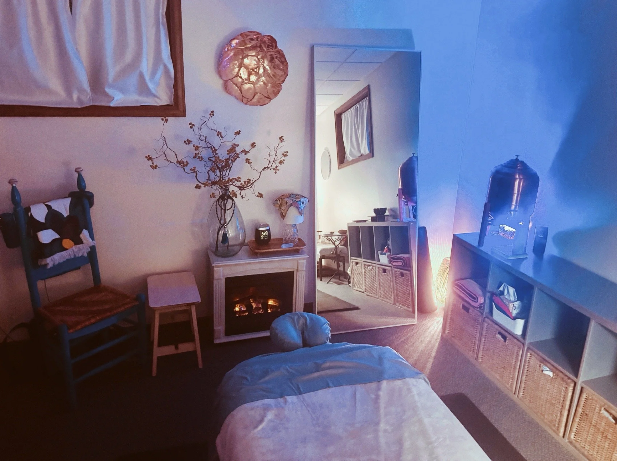 The treatment room at My Tiny Massage Studio in warm afternoon light — massage table dressed in crisp linens, small fireplace, branches in a vase